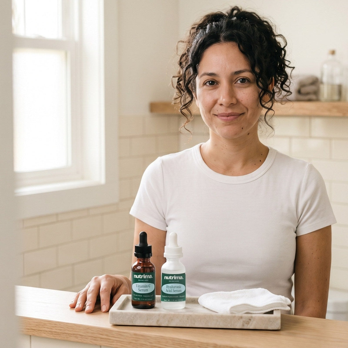 nutrima Skincare collection: Vitamin C Serum and Hyaluronic Acid Serum bottles on a bathroom vanity tray, a woman standing behind them — AI-generated brand image