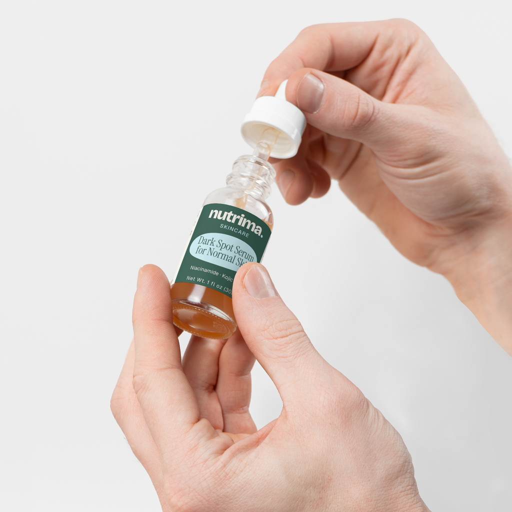 Hands opening a dropper bottle of Dark Spot Serum for Normal Skin by nutrima on a clean white background.