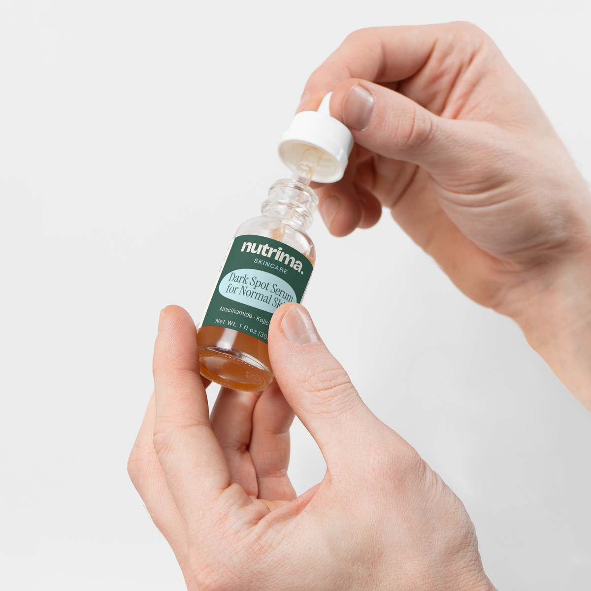 Hands opening a dropper bottle of Dark Spot Serum for Normal Skin by nutrima on a clean white background.