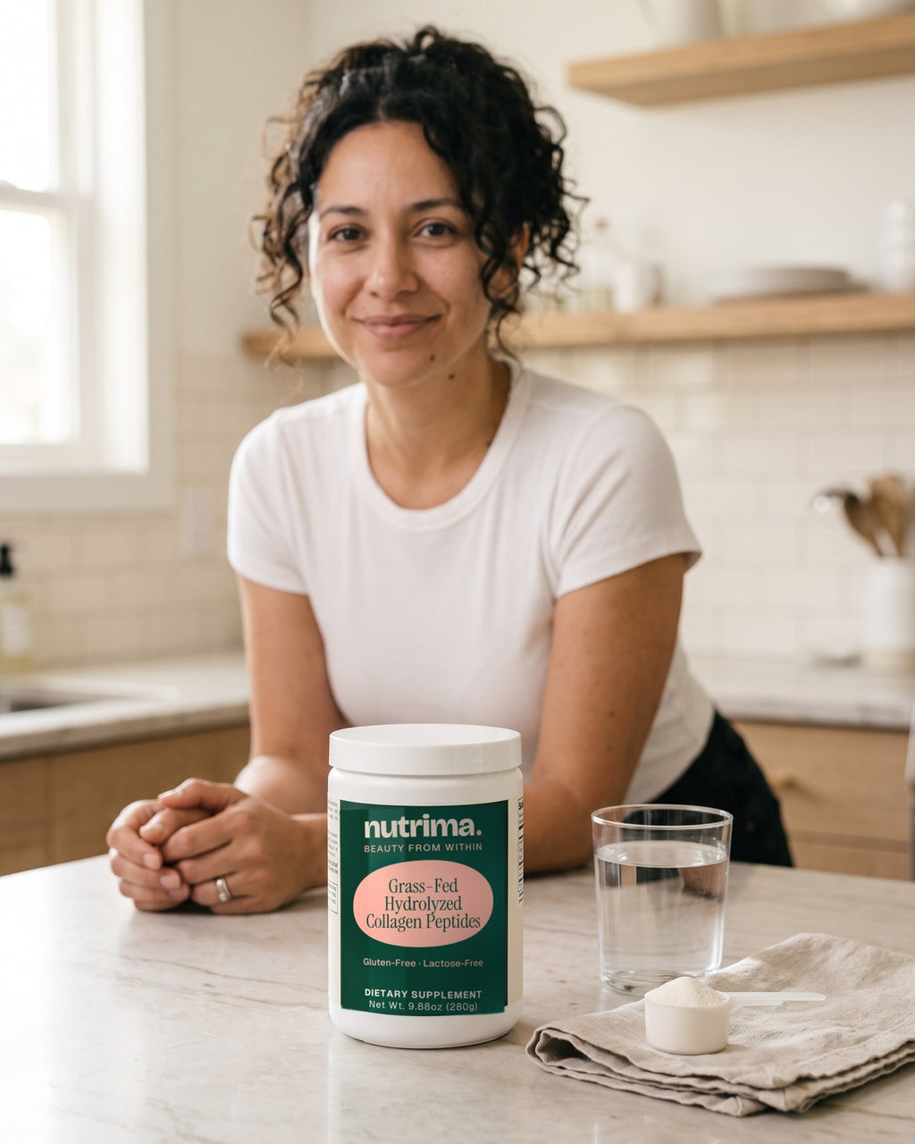 nutrima Grass-Fed Hydrolyzed Collagen Peptides tub on a marble counter beside a glass of water and a scoop of powder, a woman in the background — AI-generated brand image