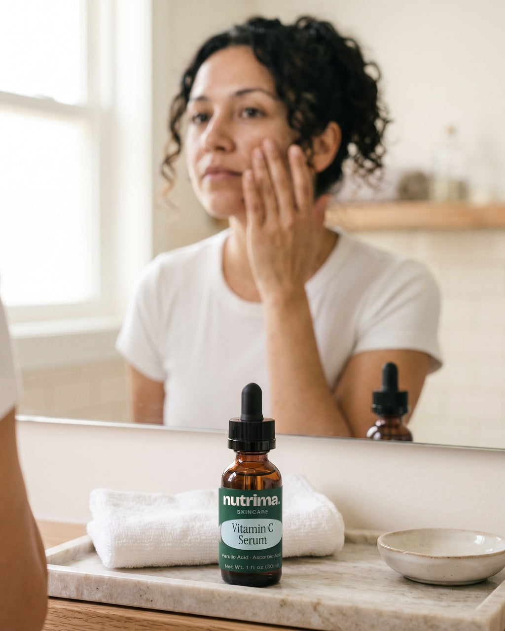 nutrima Vitamin C Serum bottle on a bathroom vanity tray, a woman at the mirror in the background — AI-generated brand image