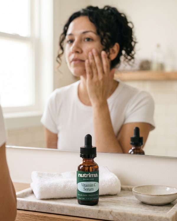 nutrima Vitamin C Serum bottle on a bathroom vanity tray, a woman at the mirror in the background — AI-generated brand image