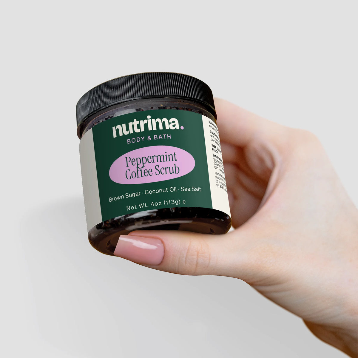 A hand holds a jar of Peppermint Coffee Scrub by nutrima, featuring a dark container and vibrant label details.